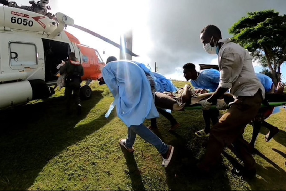 Coast Guardsmen and civilians rush toward a helicopter, carrying a patient on a stretcher.