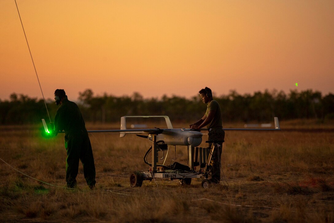 Marines work on a small unmanned aerial surveillance system illuminated by a green light under a sunlit sky.