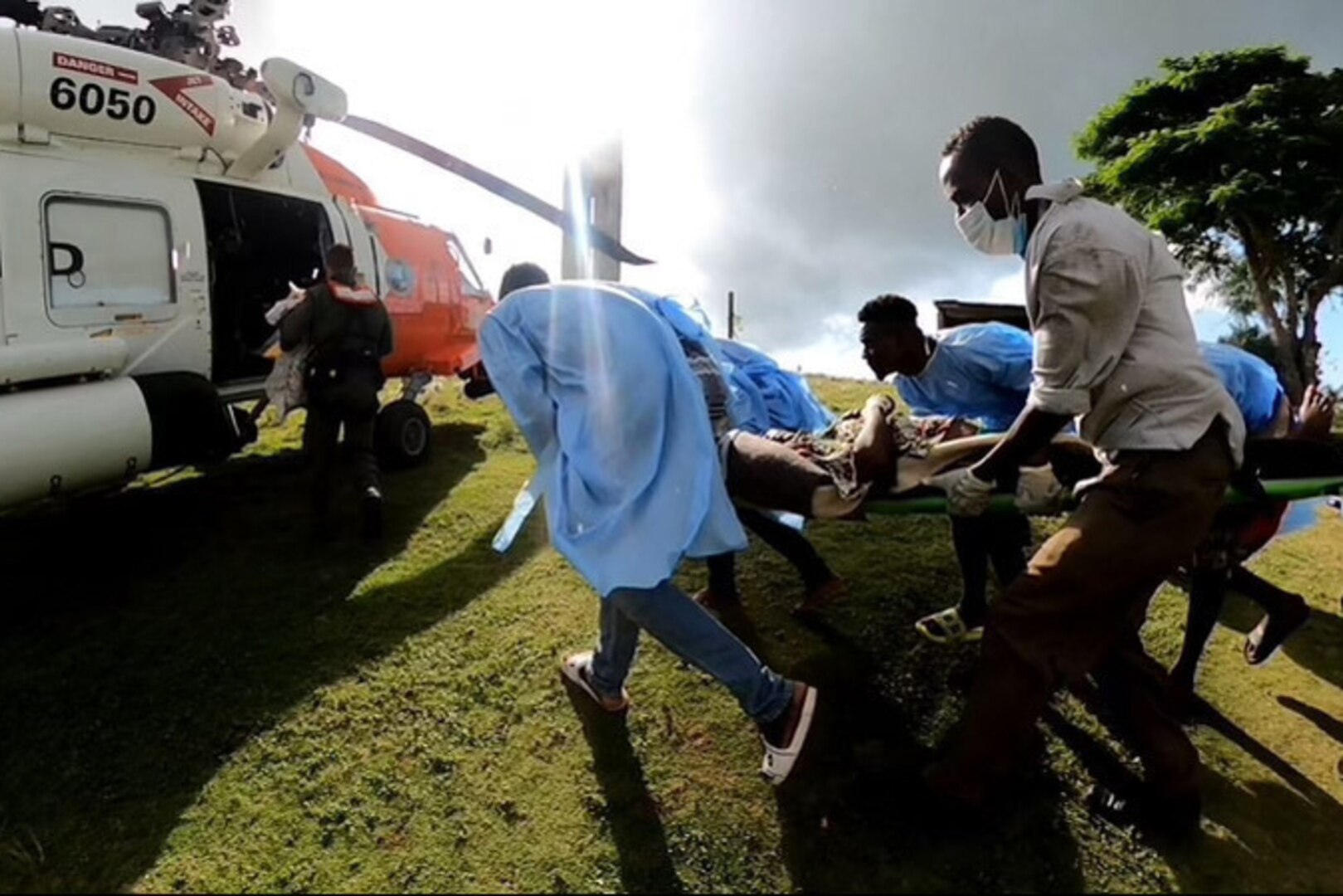 Coast Guardsmen and civilians rush toward a helicopter, carrying a patient on a stretcher.