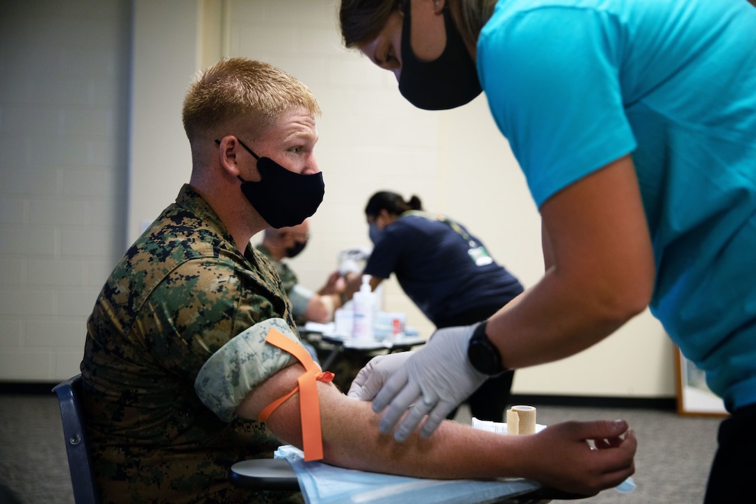 A Marine wearing a face mask sits while donating blood and a woman wearing a face mask and gloves assists.