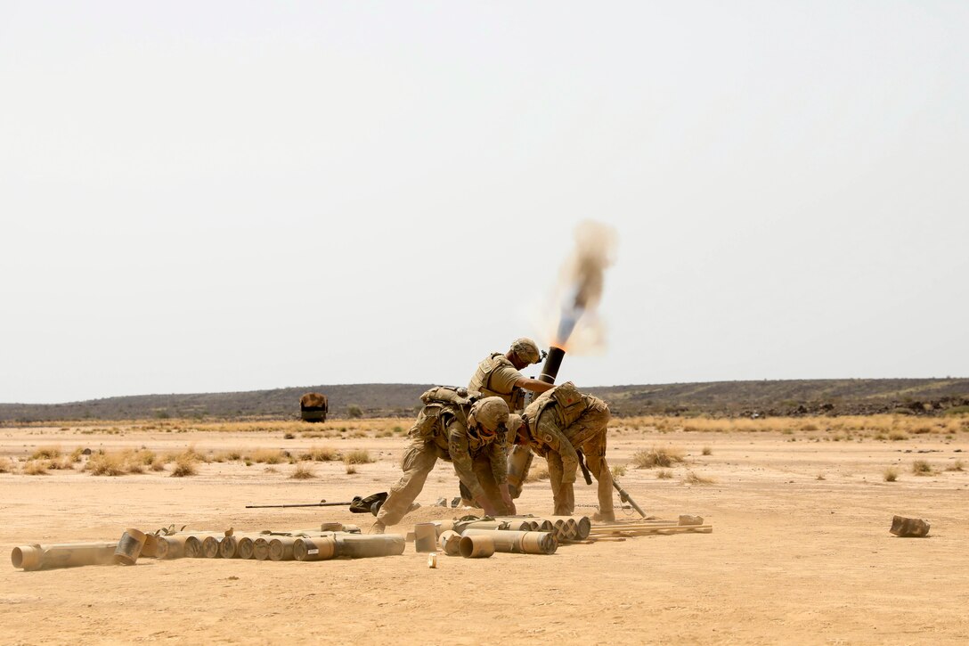 Three soldiers duck after firing a weapon from a launcher in the desert.