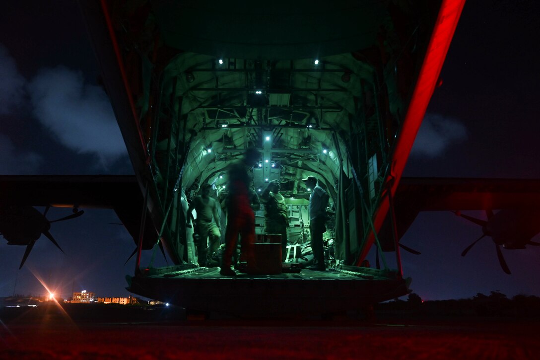Airmen stand near cargo in an open aircraft illuminated by green light.