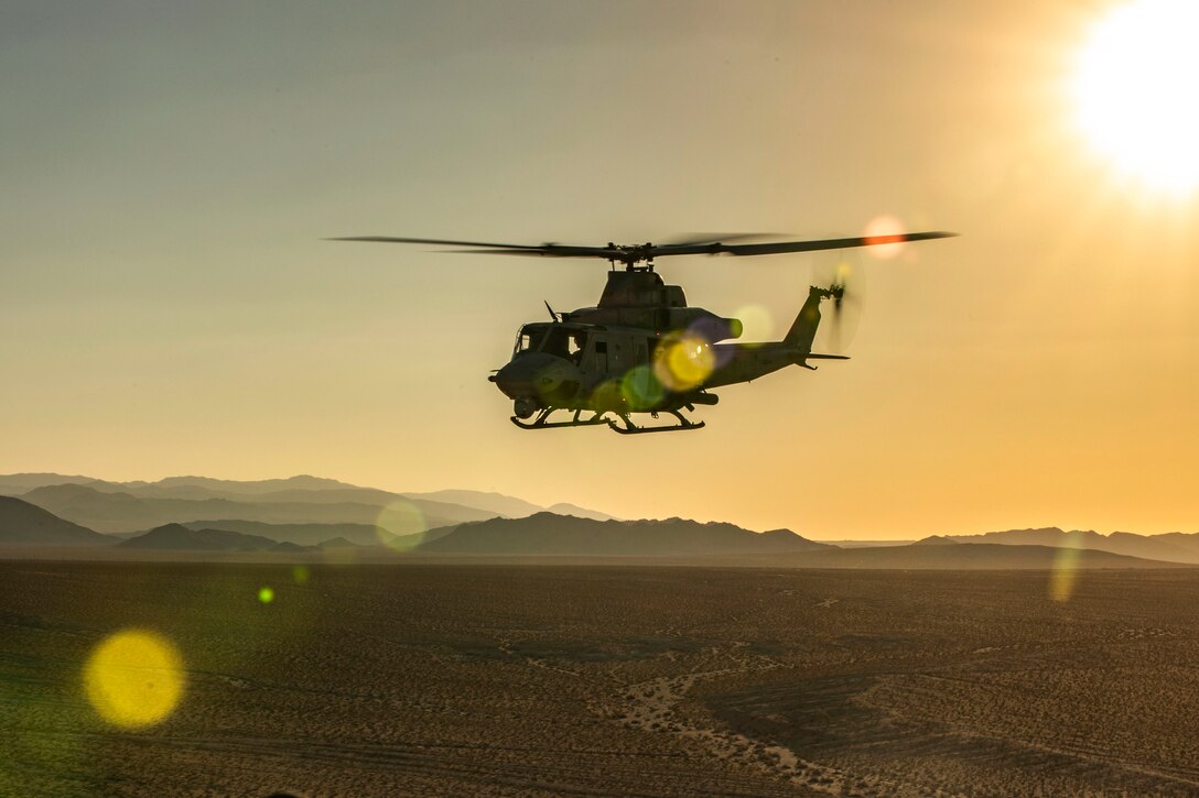 A helicopter flies over a desert under a sunlit sky.