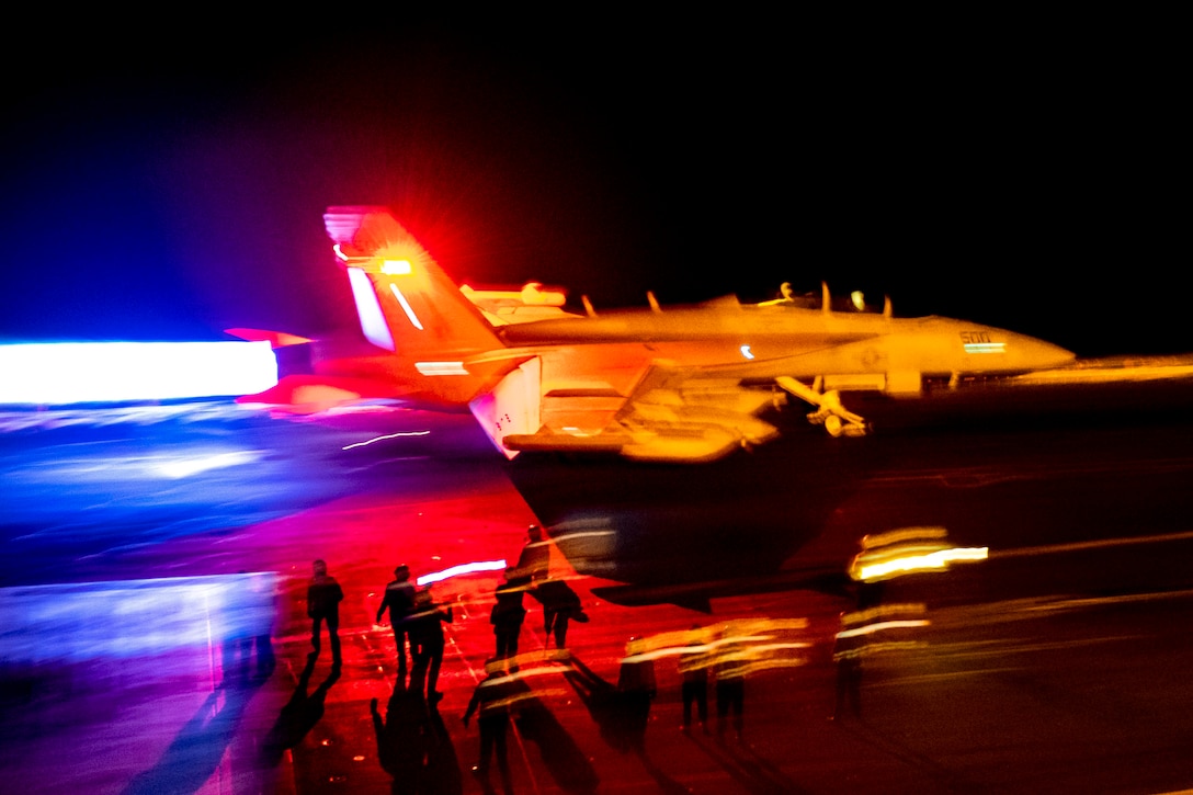 A military jet prepares to take off from the brightly lit flight deck of an aircraft carrier.