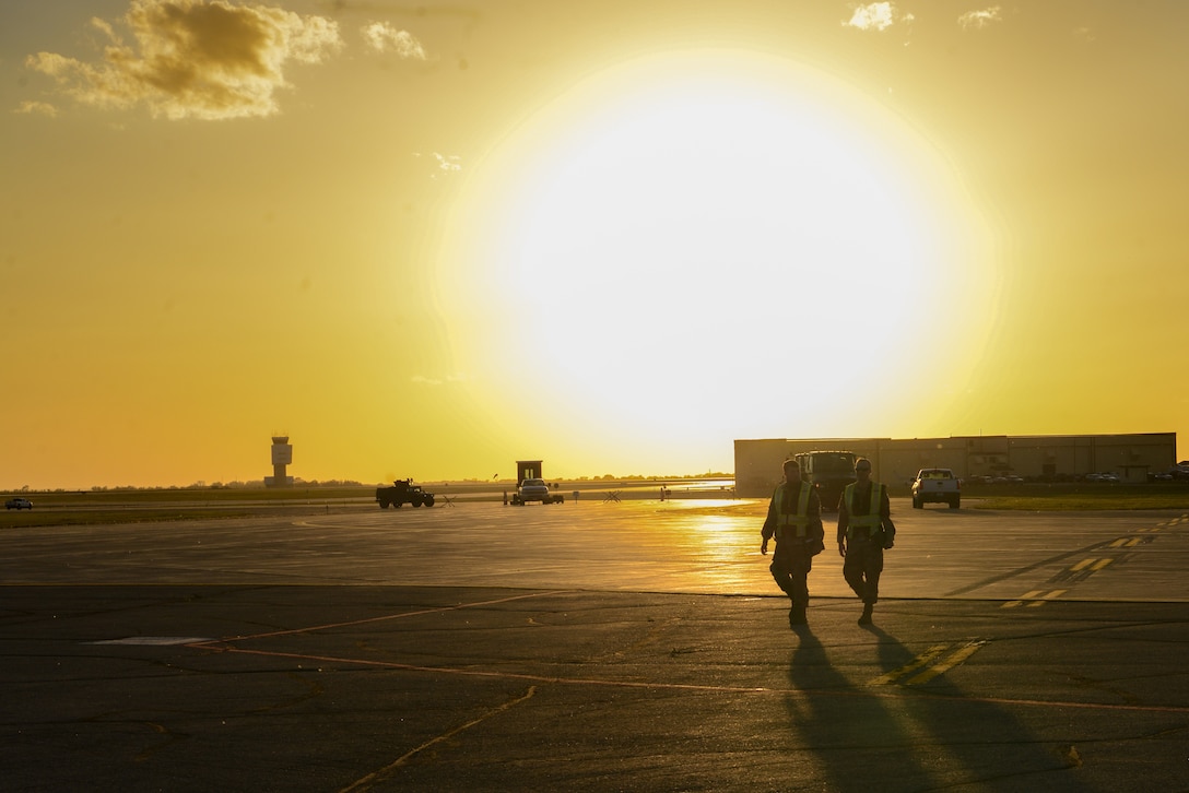 Two airmen walk along the tarmac with a large sun on the horizon.