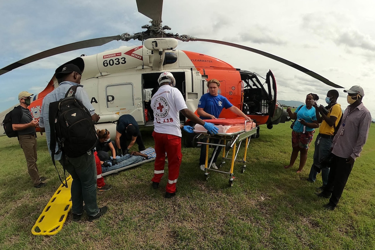 Coast Guardsmen load a patient from a stretcher onto a helicopter as other people watch.