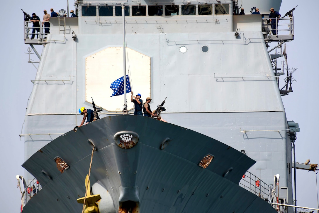 A sailor lowers a flag on the deck of a ship as others stand around.