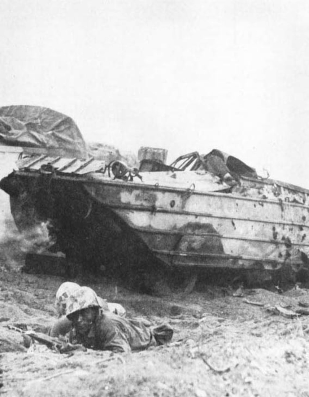Marines take cover on a beach; a battered amphibious vehicle is behind them.