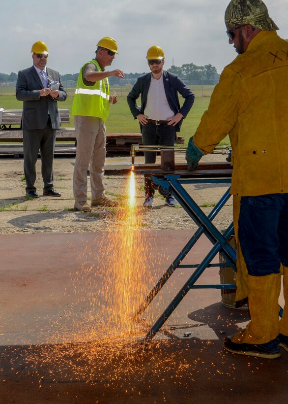 Man cuts metal using Petrogen torch while three men with safety gear watch from a distance.