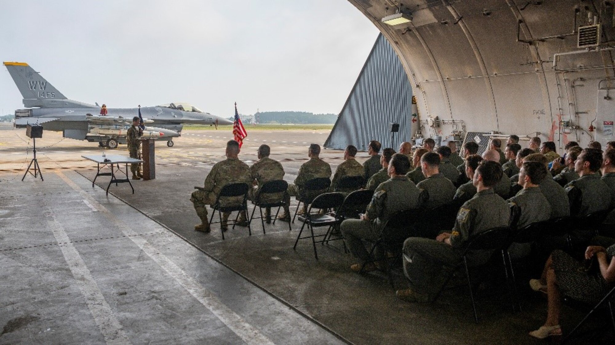 Airmen in a hanger attending a ceremony with fighter jet in the background.