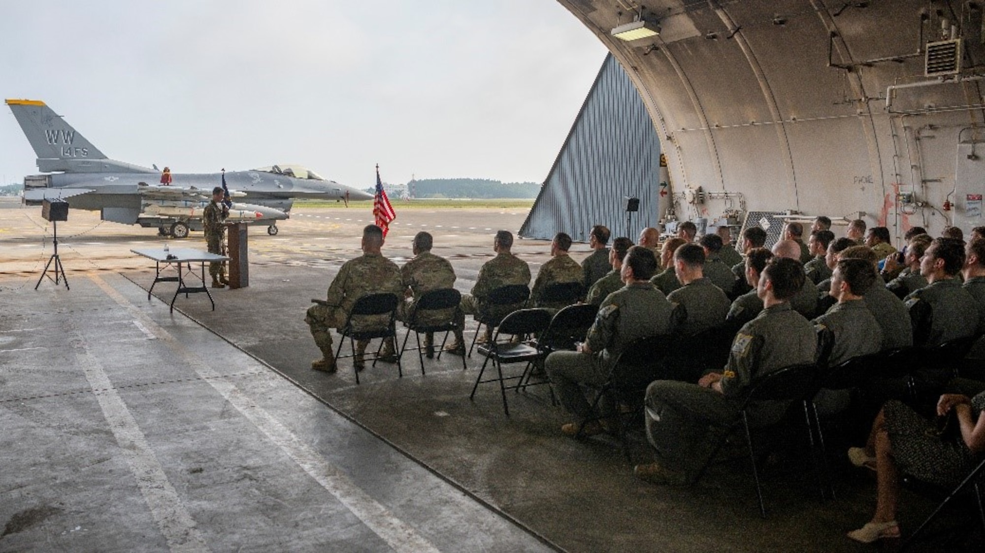 Airmen in a hanger attending a ceremony with fighter jet in the background.