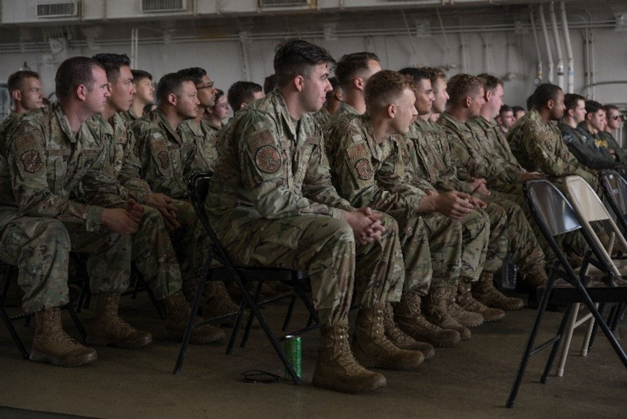 Military members sitting in a row of chairs at a ceremony in a hanger.