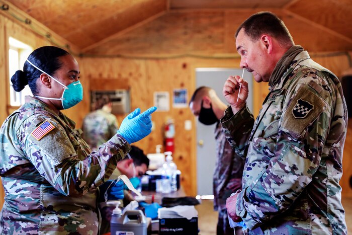 A female soldier wearing a face mask and gloves points a finger and watches as another soldier, whose face mask is pulled down beneath his chin, swabs his nose.