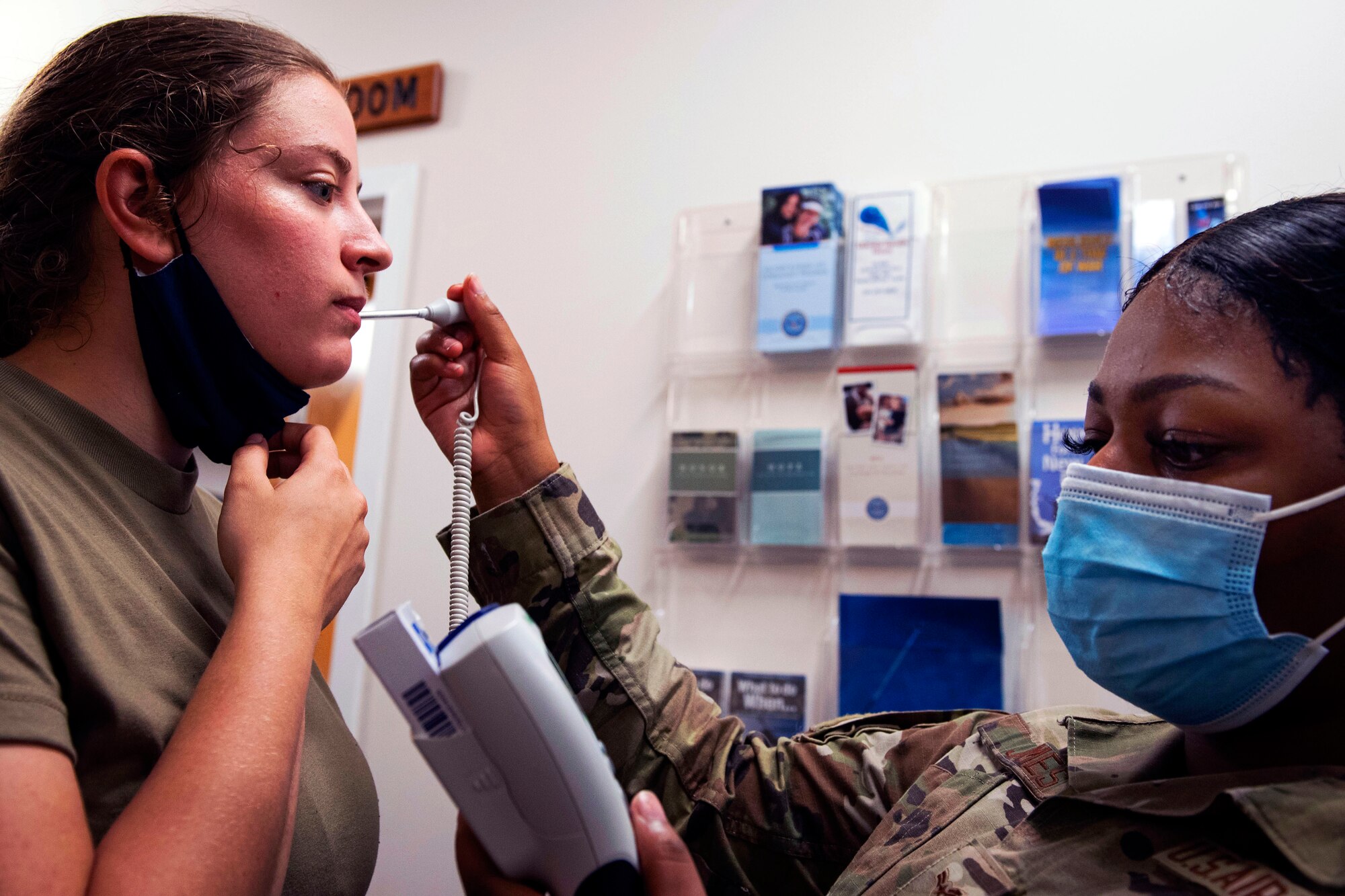 A female airman wearing a face mask takes the temperature of another  female airman, who has pulled her face mask down under her chin.