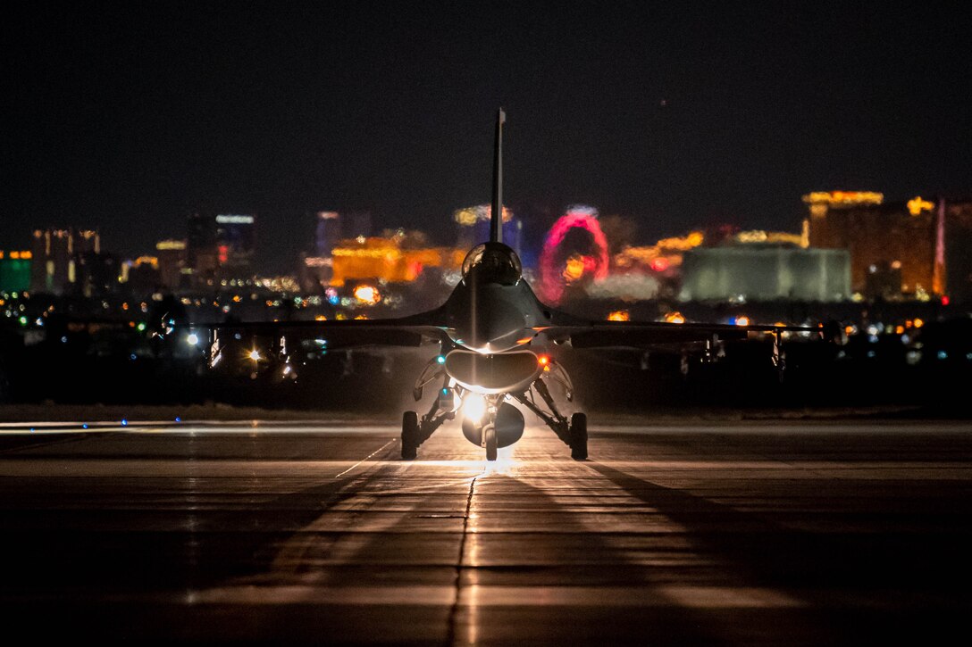A fighter jet prepares for takeoff with the lights of a city in the background.