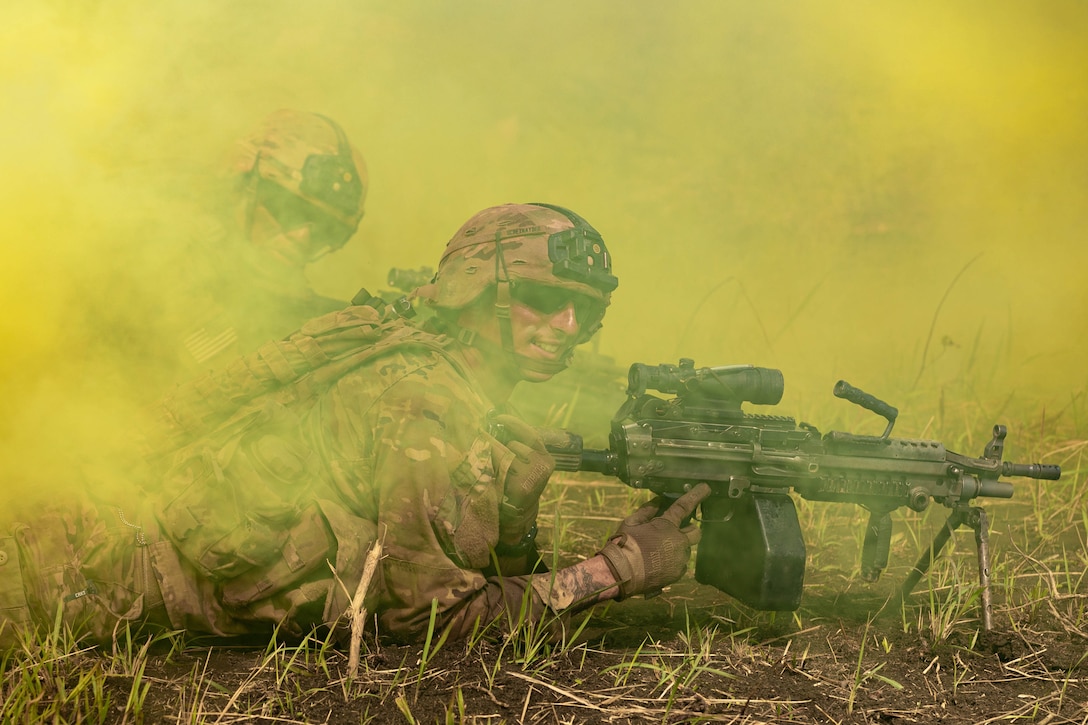 A soldier smiles as he lies on the ground holding a machine gun with yellow smoke surrounding him.
