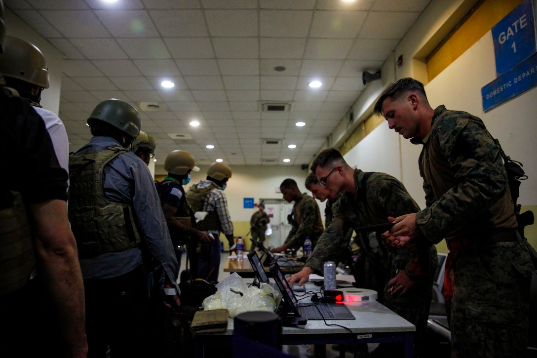 Marines stand behind a table with laptops and speak to personnel standing in a line wearing security gear.