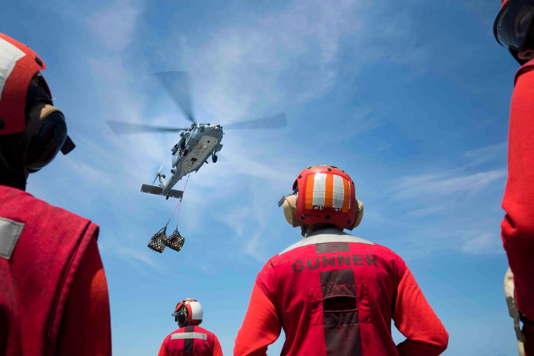 Sailors look up toward an airborne helicopter.