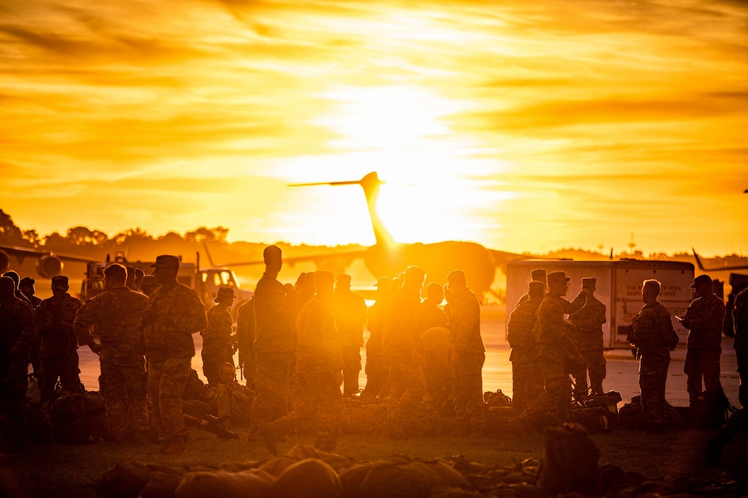 Soldiers stand as a group as the sun shines behind parked aircraft.