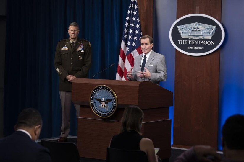A man in a suit stands at a podium addressing an audience, an Army officer stands behind him.