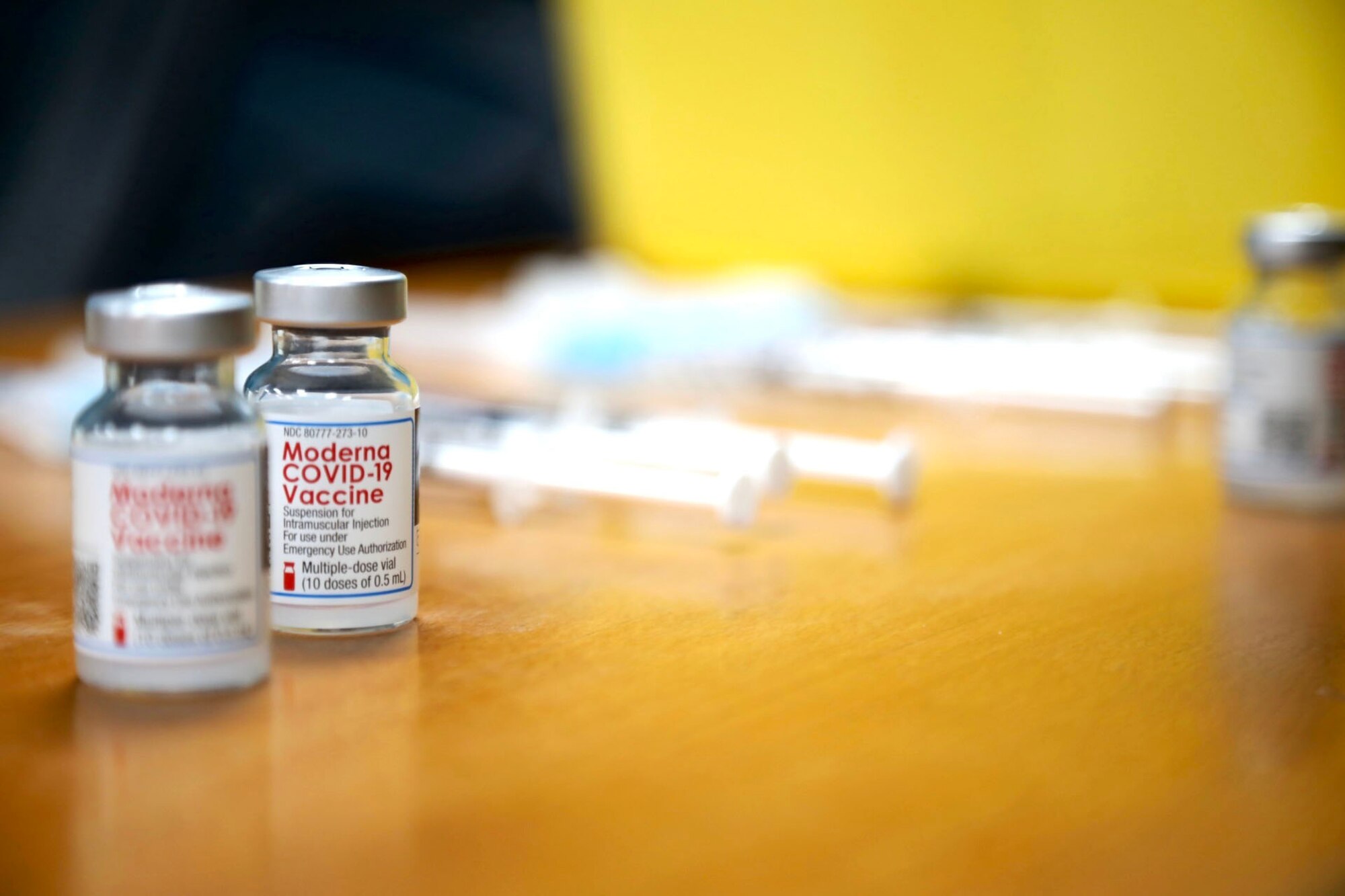 Several small vaccine bottles sit on a table along with several syringes and another small collection of vaccines in the background.