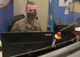 Army Reserve Spc. Noah Smock, an information technology specialist, or 25 Bravo, mans the computer and telecommunications Help Desk at 1st Theater Sustainment Command's operational command post, wearing his face mask and behind a plastic shield--all part of the 1st TSC-OCP's COVID-19 mitigation protocols. Smock deployed to the 1st TSC-OCP at Camp Arifjan, Kuwait, with other "Brickyard" Soldiers of the Indianapolis-based 310th Sustainment Command (Expeditionary), who staff the 1st TSC-OCP during their nine-month rotation that began December 2020.