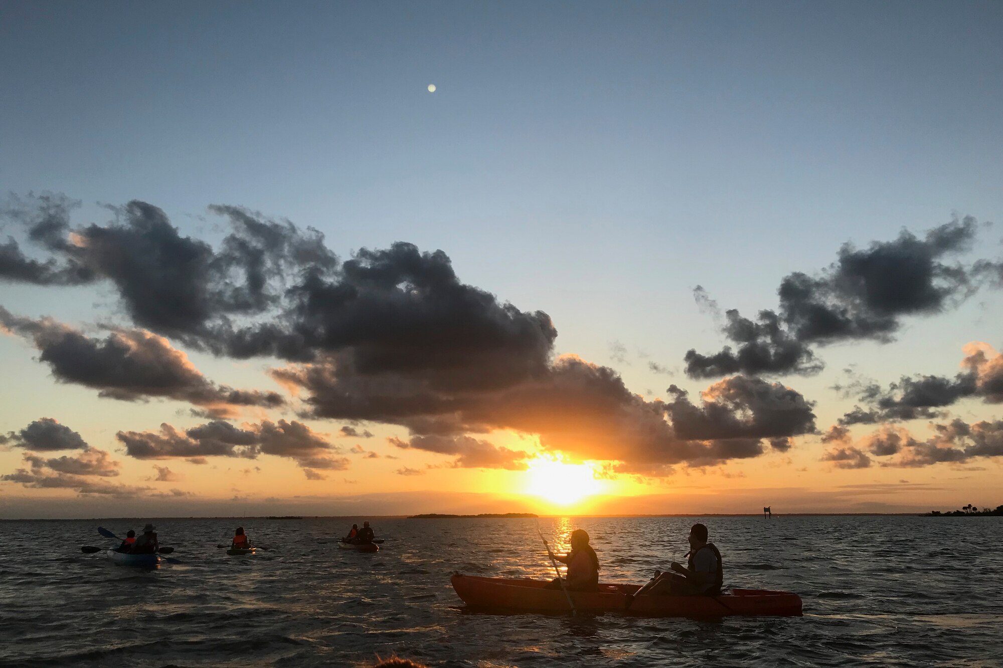 A photo of people sitting in kayaks on the water in front of a sunset.