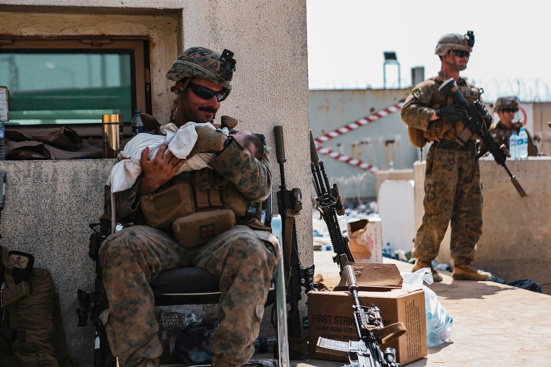 A Marine sits and holds a baby while another stands watch.