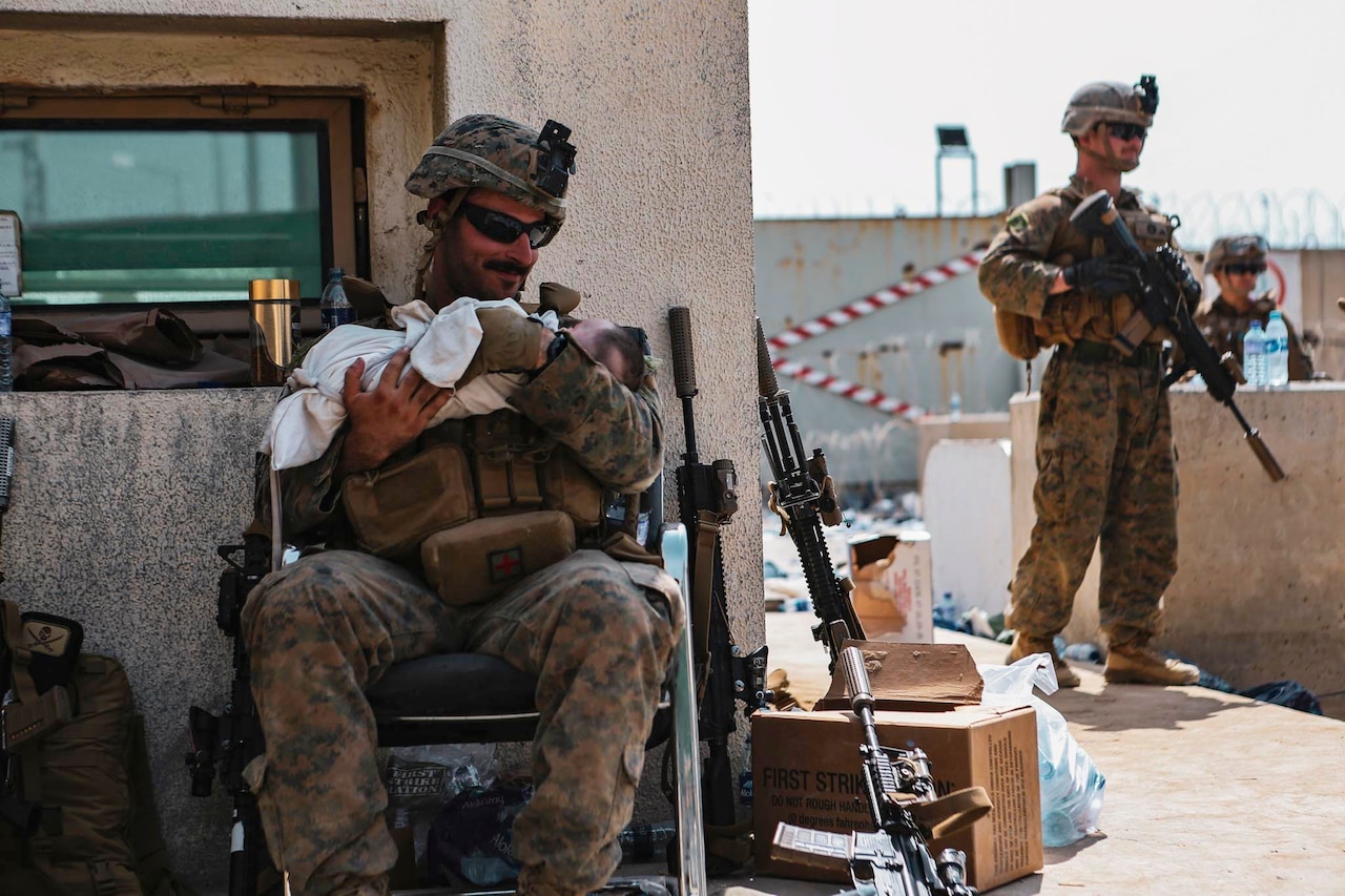 A Marine sits and holds a baby while another stands watch.