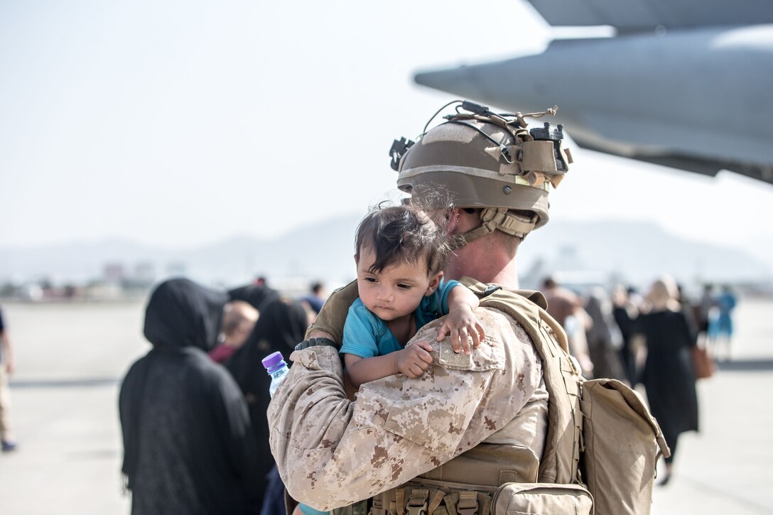 A Marine holds an infant on his shoulder while others line up to board an aircraft.