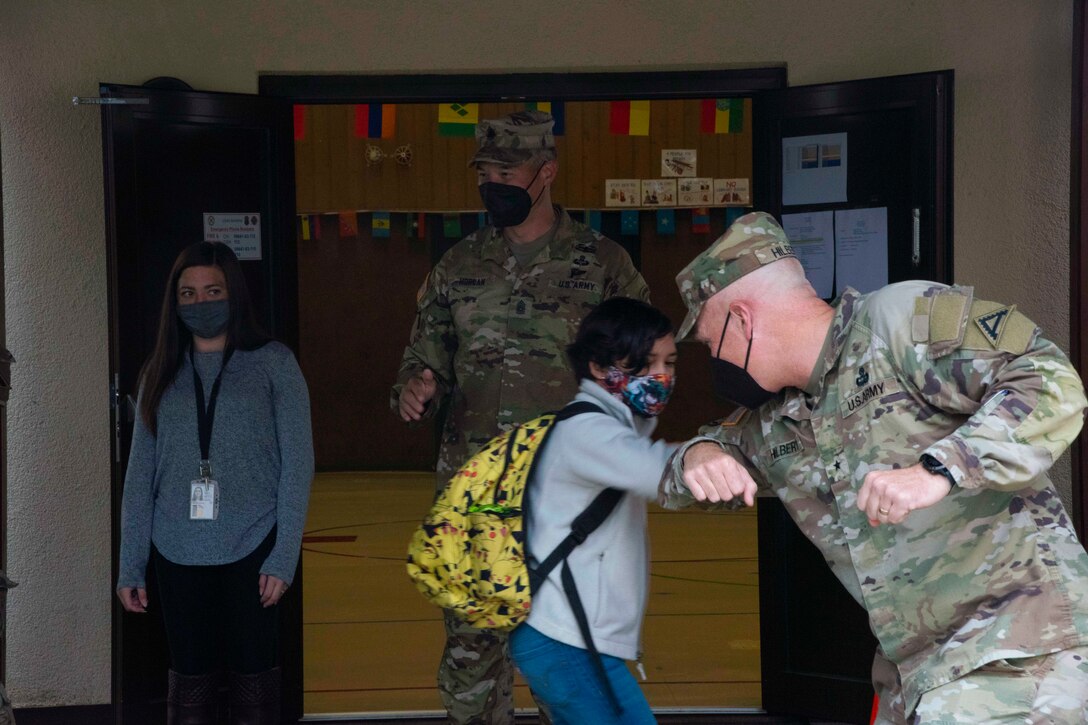 A soldier bumps elbows with a student as a fellow soldier and teacher watches outside of a school building.