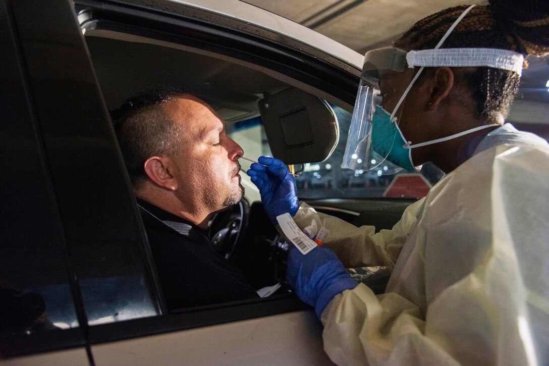 A female medical technician wearing personal protective equipment inserts a nasal swab into the nostril of a man sitting in his vehicle for a COVID-19 test.