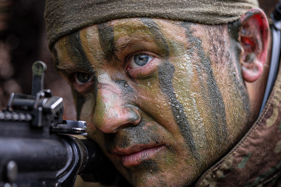 A closeup of a soldier wearing  face paint  as he aims his weapon.