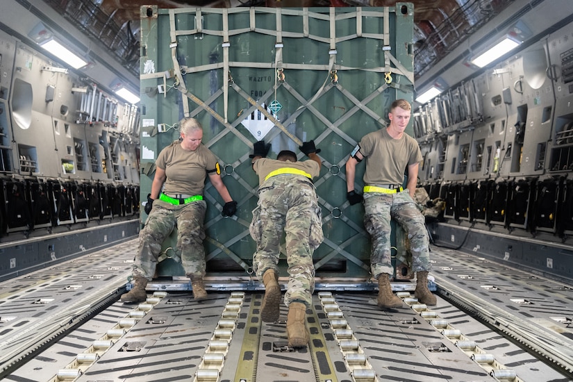 Three airmen push  a large load of supplies on an aircraft.