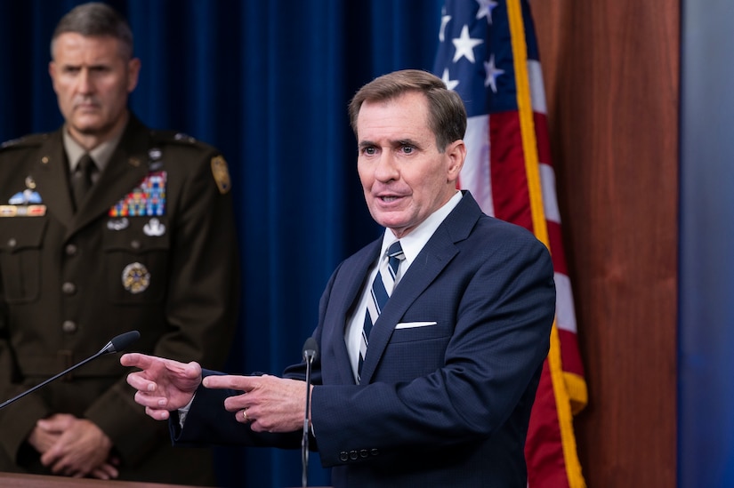 A man dressed in a business suit appears to speak and point to his right; a man in a military uniform stands in the background.
