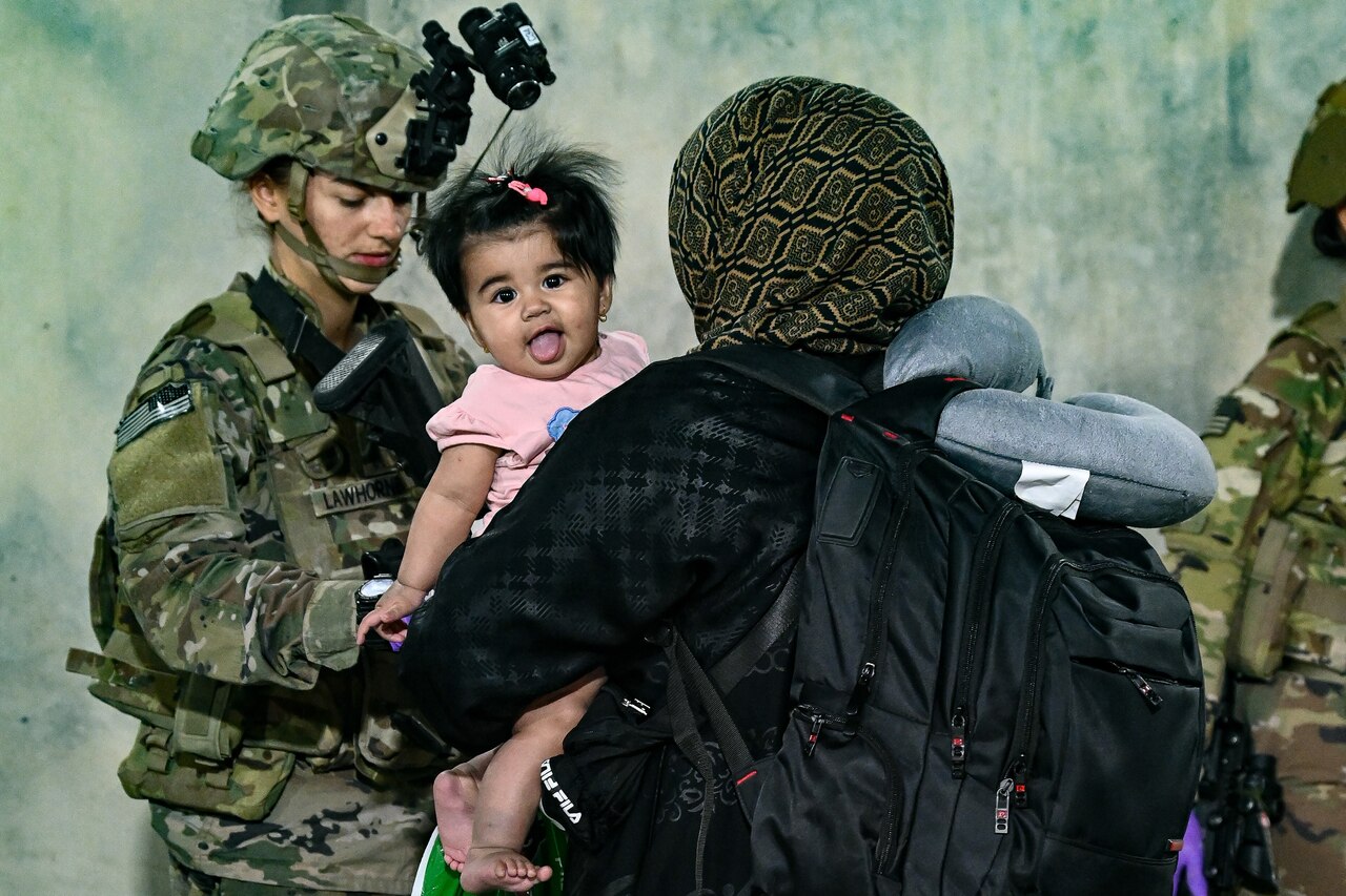 A woman holds a baby.  A U.S. service member stands near the woman.