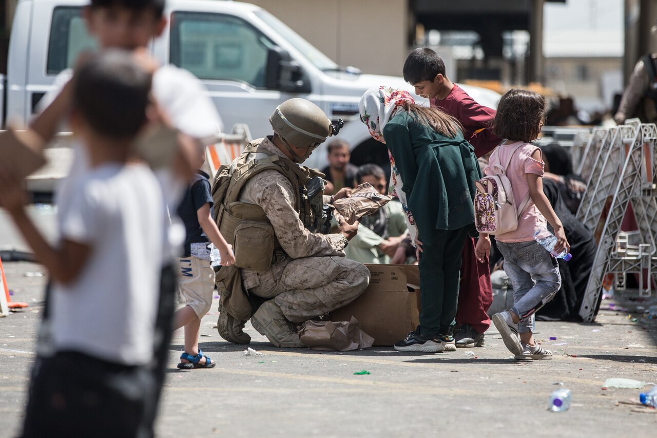 A U.S. service member kneels on the ground. Children stand around him.