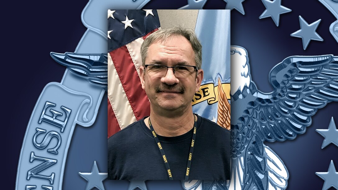Head and shoulders image of white guy wearing glasses in front of the US and  DLA flags.