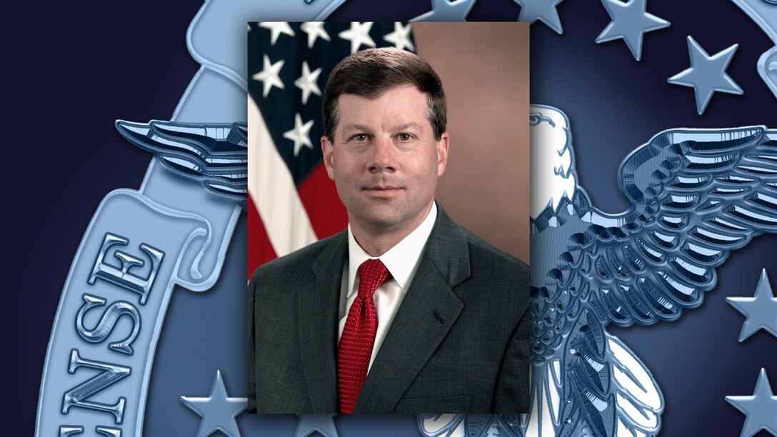 White man in a black suit and red tie sits in front of the US flag.