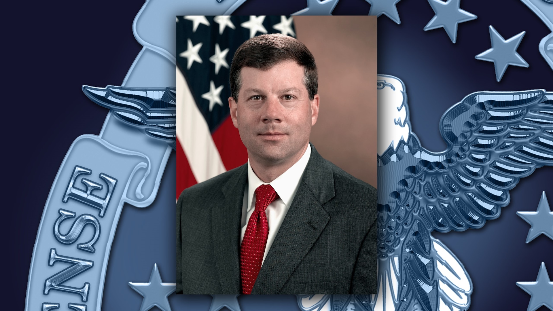 White man in a black suit and red tie sits in front of the US flag.