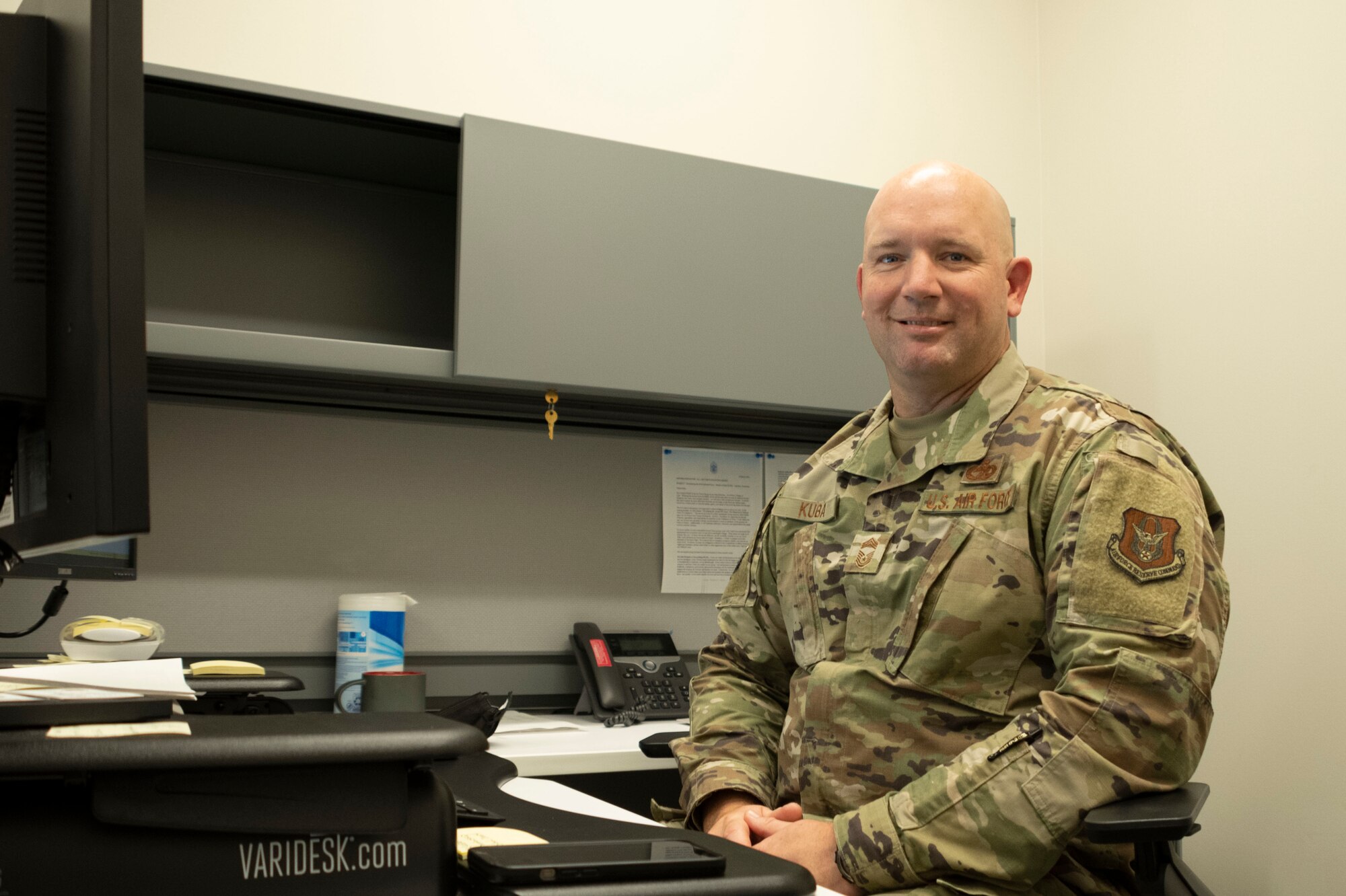 A man smiles behind a computer desk.