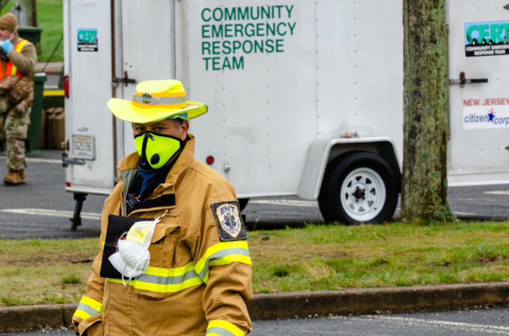 An emergency responder in a reflective yellow coat stands in the foreground in a parking lot with a trailer in the background labeled "Community Emergency Response Team."