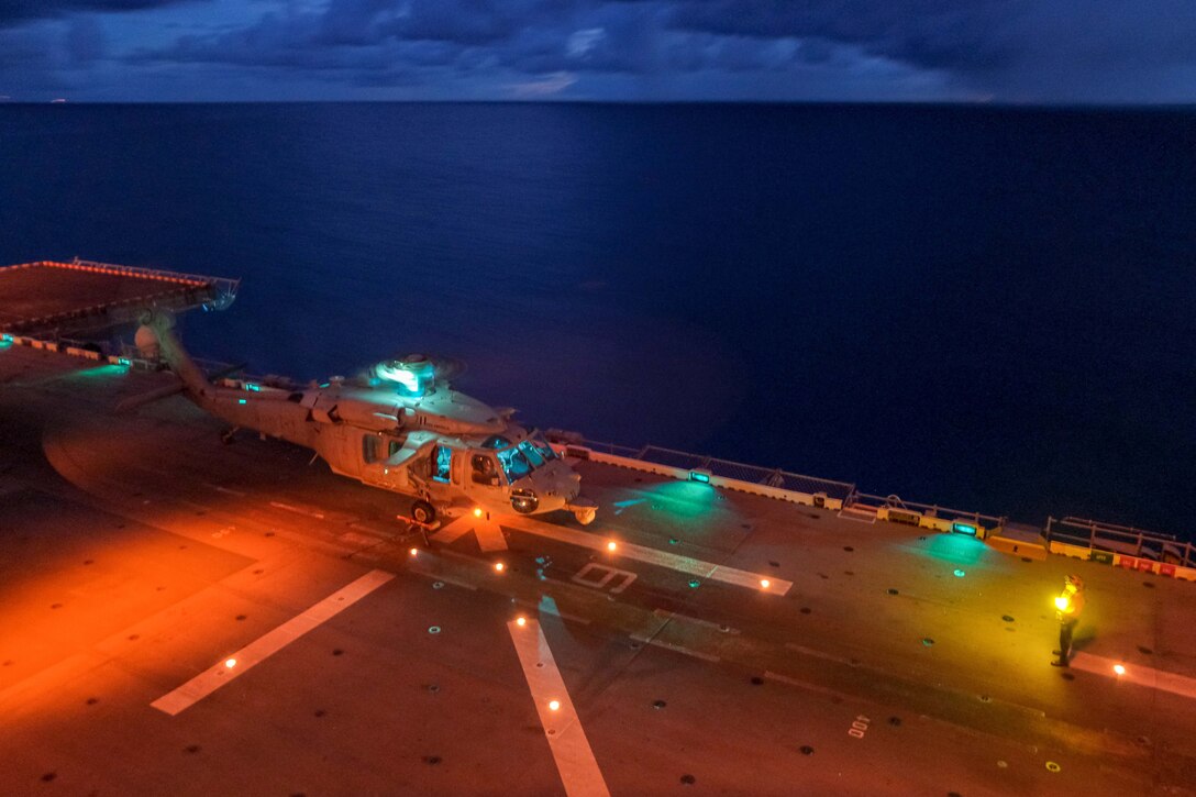 A helicopter prepares to take off from a ship at sea illuminated by colorful lights.