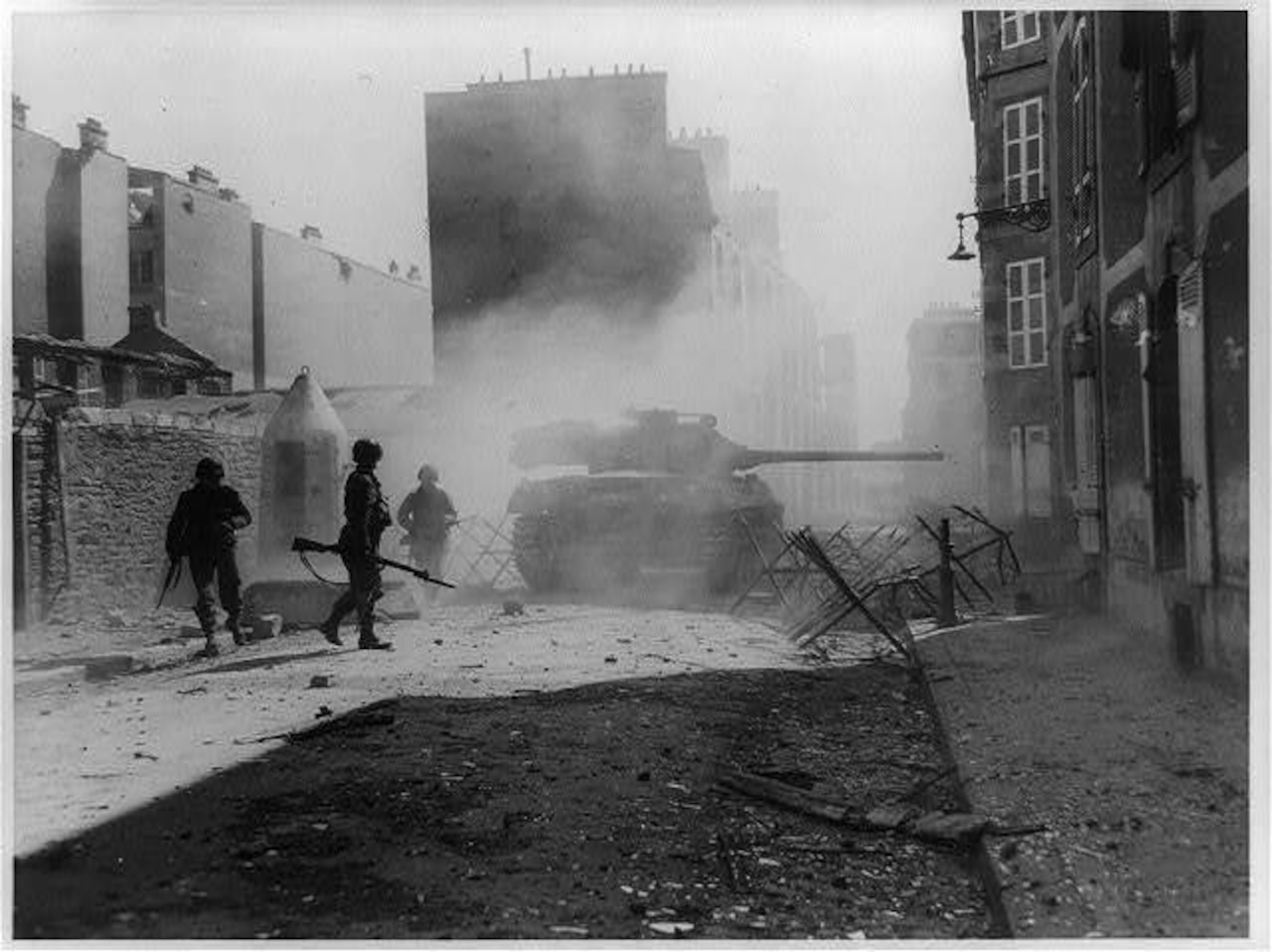 Three soldiers with guns stand near a tank on an empty, debris-covered street.