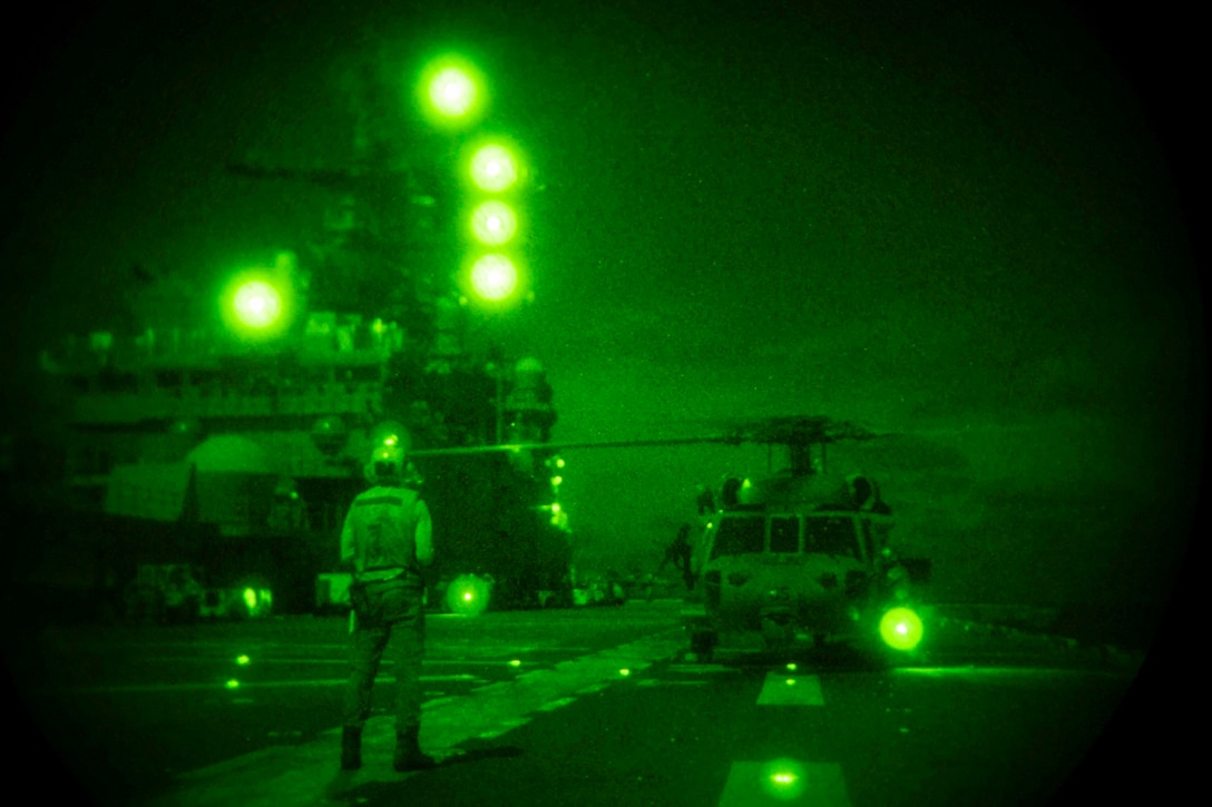 A sailor illuminated by green light stands in front of a helicopter on the deck of a ship at night.