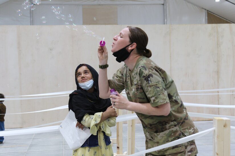 A U.S. Army National Guard Soldier with Task Force Spartan, U.S. Army Central, entertains an Afghan girl with bubbles as she waits to in-process at Camp Buehring, Kuwait, Aug. 23, 2021. U.S. Army Soldiers continue to work with their U.S. Central Command and Department of State teammates to support Afghanistan evacuation efforts with transportation, security, logistics and medical assistance at locations in Kuwait. (U.S. Army Photo by 1st Lt.  James Mason)