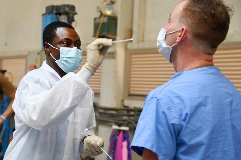 A man wearing a face mask and gloves holds a nasal swab while another man wearing a face mask tilts his head back.