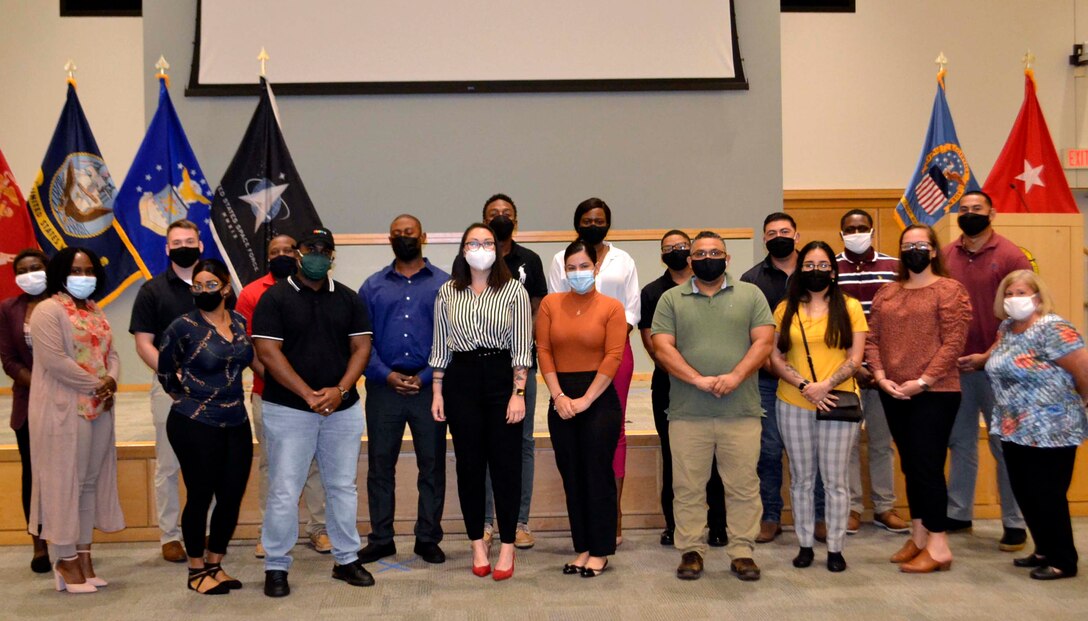 A group of soldiers dressed in civilian business casual attire stand in front of a low stage adorned with flags of the U.S., Defense Logistics Agency and military services.