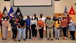 A group of soldiers dressed in civilian business casual attire stand in front of a low stage adorned with flags of the U.S., Defense Logistics Agency and military services.