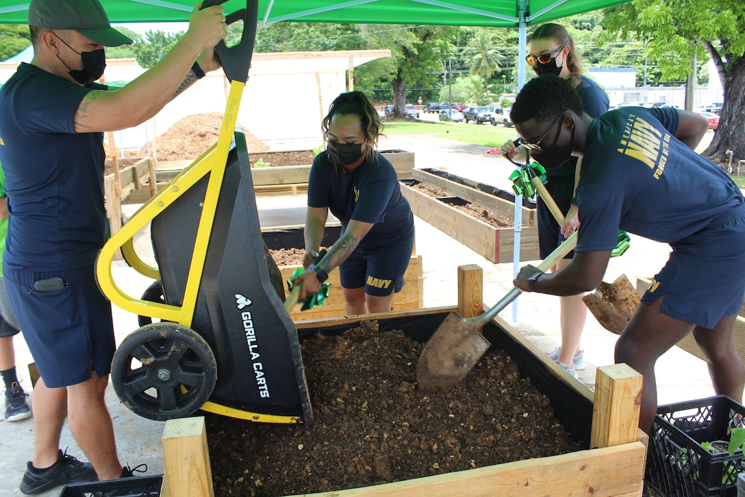A group of service members use a wheelbarrow and tools to prepare a garden bed.